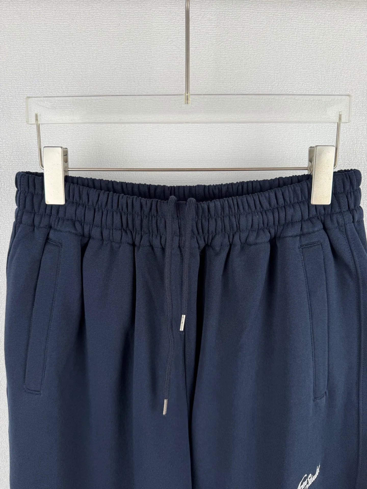 ACNE STUDIOS embroidered logo fleece sweatpants wide-leg trousers. Fabric: 32-count double yarn top + 16-count terry bottom yarn G The veil is made of 32-count fine yarn. The high-count yarn is smooth and delicate. After weaving, it forms a smooth and compact surface. It not only resists pilling and wear-resistant, but also gives the fabric a touch as delicate as skin. The base yarn is made of 16-count roving. The low-count yarn is fluffy and hairy. It naturally forms a rich terry layer during the weaving process, providing a sufficient source of fiber for subsequent sanding. This combination of fine surfaces and thick soles strikes a perfect balance between warmth and skin-friendliness. Mechanical sanding is the soul process of this fabric. By rubbing the bottom surface of the roller wrapped with emery leather at high speed, the fiber ends of the 16-count roving are evenly pulled out and cut off, forming a dense and uniform short pile layer. The smoothness and neatness of needle embroidery and the soft velvet feel of brushed fabrics form a material dialogue between hardness and softness. The embroidery area is smooth and crisp, with sharp lines, like delicate inscriptions carved on a rich suede canvas; while the surrounding brushed fabric presents a soft matte natural texture. The spacing between stitches is consistent and the strength is balanced to ensure that each stitch is placed accurately. This rigorous stitching structure makes the edges of the logo present a sharp and clear outline, and the strokes of the text are full and smooth. Even the curved turning points of the floral fonts can achieve a smooth transition through the connection of tight stitches.